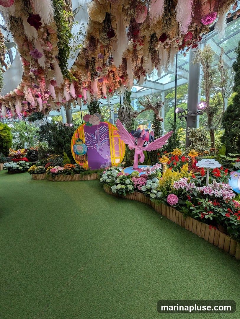 A vibrant indoor garden display featuring a lush green carpeted floor, surrounded by a variety of colorful flowers and plants. Hanging floral arrangements adorn the ceiling. In the center, a cutout of a traditional Chinese-style circular window with fireworks and a lantern is visible, alongside a pink My Little Pony character figurine with wings. Small signs with text are placed among the flowers.