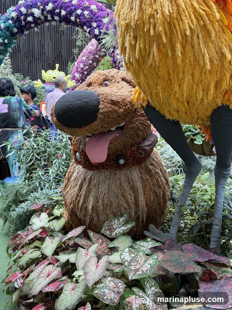 A whimsical floral display features a large, fluffy brown dog character with its tongue out, reminiscent of Dug from the movie 'Up'. To its right, a tall, yellow, feathery bird-like figure stands on thin legs. In the background, a purple and white floral arch frames a green alien character from Toy Story. People are visible in the background, observing the display. The foreground is filled with lush green and pink foliage.