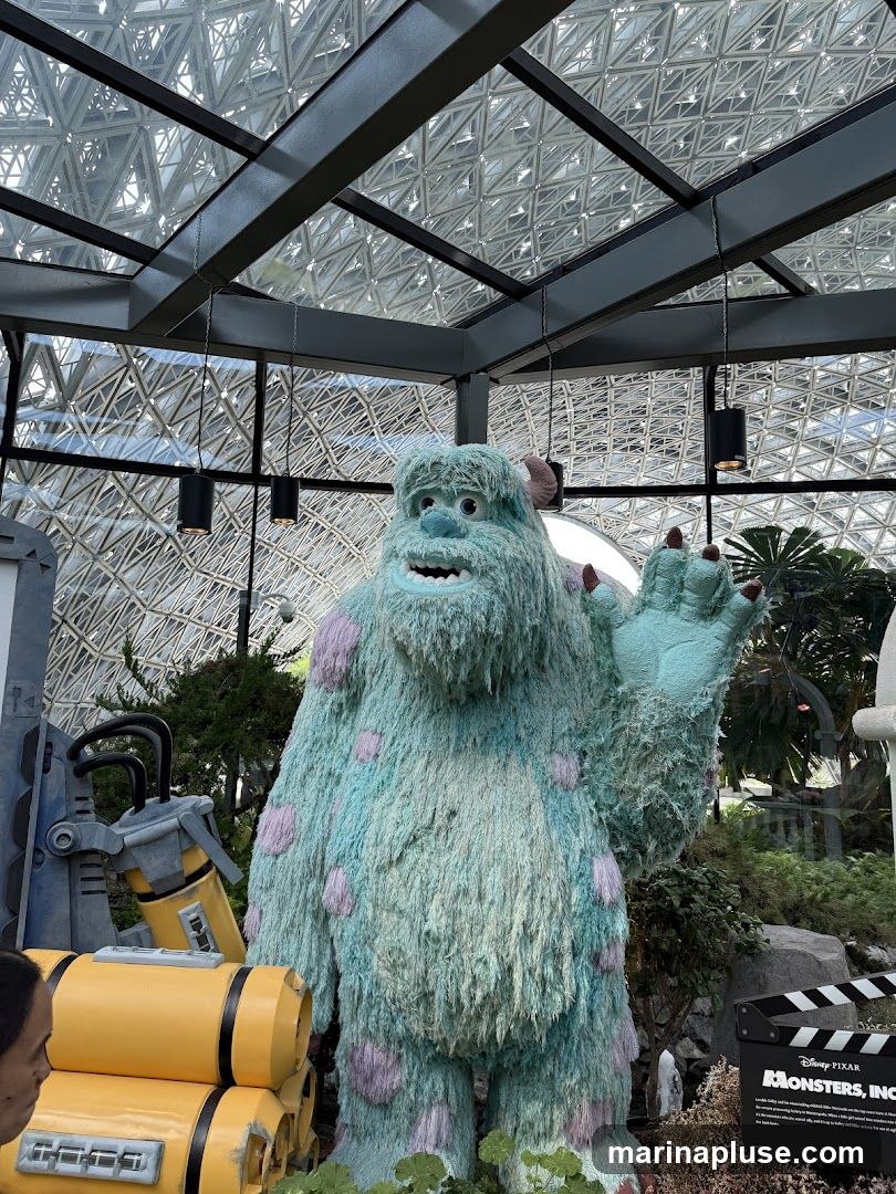 A large, fuzzy blue and purple monster statue, resembling Sulley from Monsters, Inc., stands in an indoor garden setting with a geodesic dome ceiling. To the left, yellow cylindrical objects are stacked, and to the right, a clapperboard with the "Monsters, Inc." logo is visible.