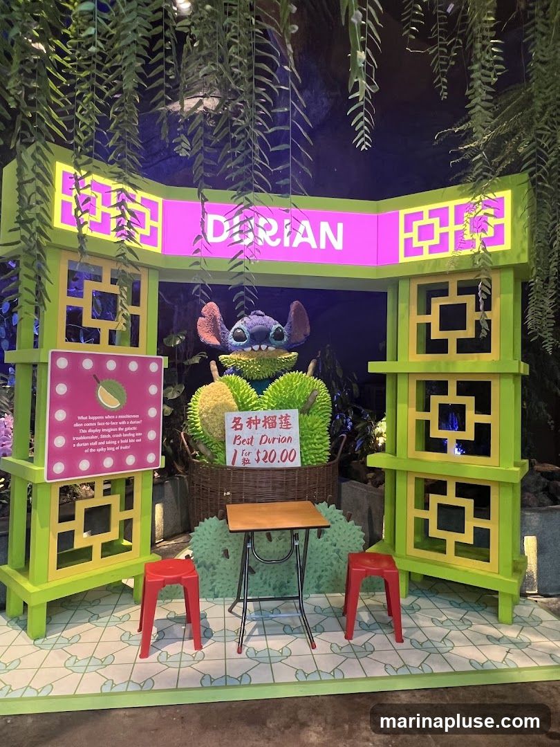 A vibrant display featuring the word "DURIAN" in large neon letters above a basket of durians. A Stitch character is placed in front of the durians. To the left, a pink sign with text and an image of a durian is displayed. To the right, a sign with Chinese characters and English text reads "Best Durian 1 for $20.00". A small table and two red stools are set up in front of the display. Green foliage hangs from above, framing the scene.