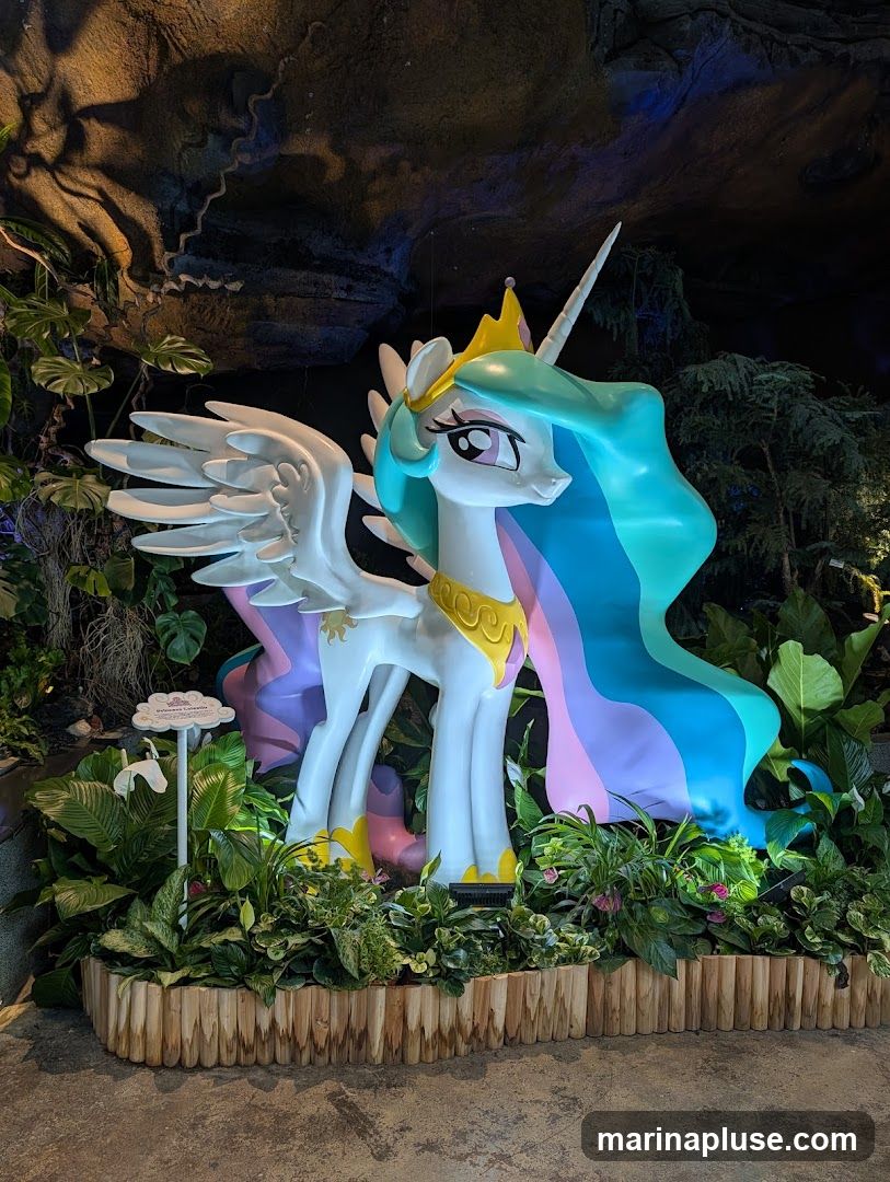 A large statue of Princess Celestia from My Little Pony stands amidst lush greenery and artificial rock formations. The statue is white with a golden crown and collar, and has a flowing mane and tail in pastel shades of blue, pink, and purple. A small sign with a cloud shape is visible to the left of the statue. The background suggests an indoor, themed environment.
