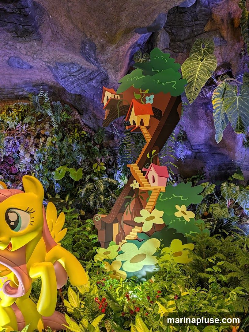 A vibrant, whimsical display featuring a large yellow My Little Pony figurine in the foreground, surrounded by lush greenery and artificial plants. Behind the figurine, a colorful, cut-out treehouse with small houses and a winding staircase is mounted against a backdrop of textured rock formations illuminated with purple and blue lights. The scene evokes a magical, enchanted forest setting, likely part of an indoor attraction or themed environment.