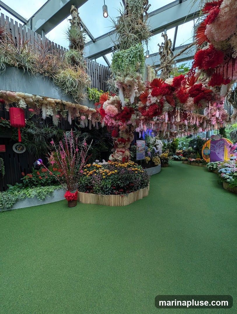 A lush indoor garden display with a variety of plants and floral arrangements. Green artificial turf covers the ground, leading to pathways lined with colorful flowers and decorative elements. Hanging plants and elaborate floral installations adorn the ceiling and walls, creating a vibrant and immersive atmosphere. A red lantern and a circular emblem are visible on the left side, suggesting a festive or cultural theme.