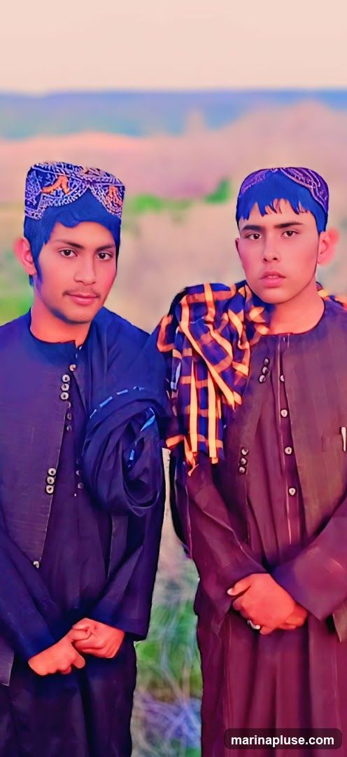 Two young men are posing for a photo outdoors. They are both wearing dark-colored, traditional outfits and embroidered caps. The man on the left has his hands clasped in front of him, while the man on the right has a patterned scarf draped over his shoulder. The background is blurred, showing a landscape with muted colors.