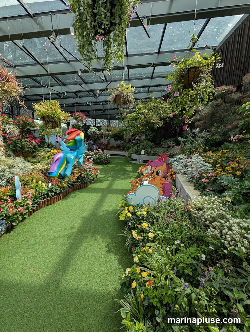 An indoor garden with a glass ceiling, featuring lush greenery, colorful flower beds, and hanging plants. A pathway made of artificial grass winds through the display. Two cartoon character figures, resembling ponies with rainbow manes, are placed among the flowers. Several people are visible in the background, suggesting it is a public space or attraction.