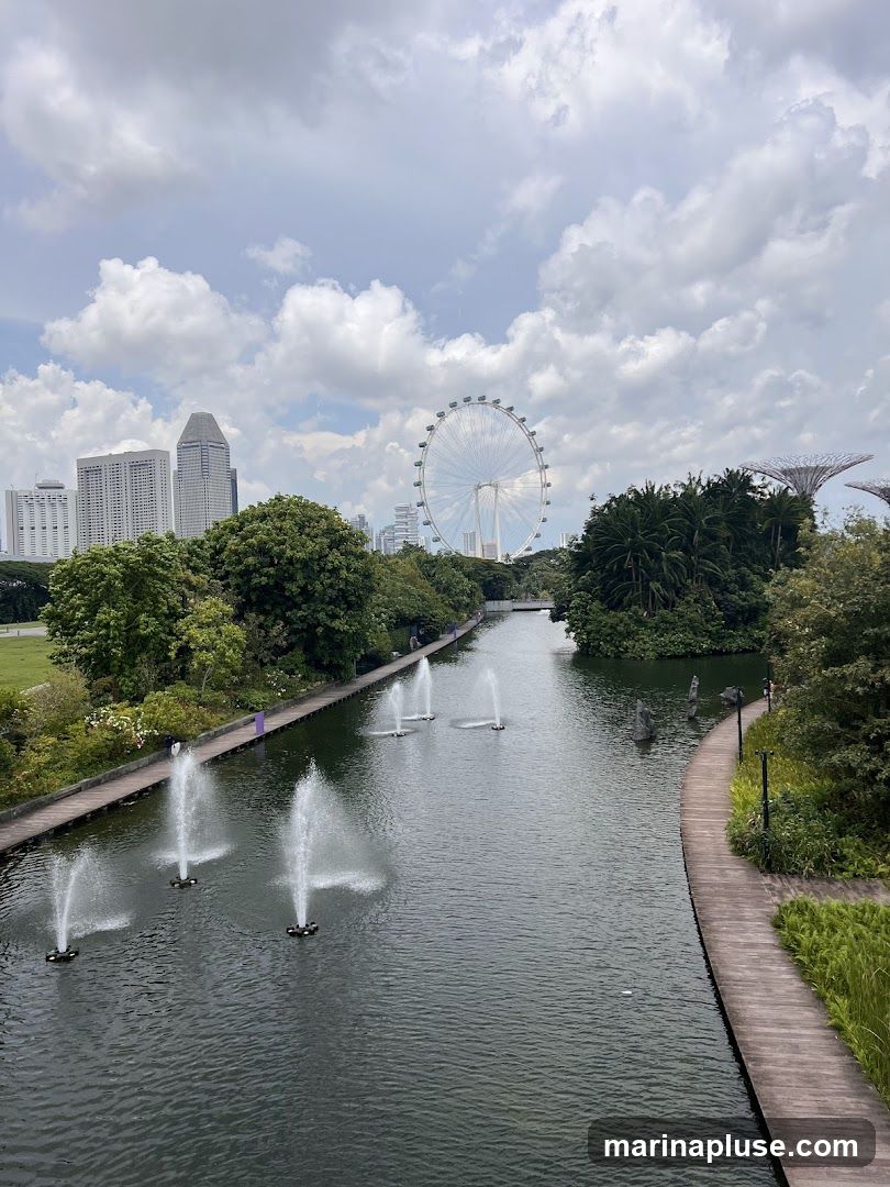 A scenic view of a park with a canal and fountains, set against a backdrop of a city skyline and a large Ferris wheel. Lush greenery and trees line the canal, with a wooden walkway on the right. The sky is partly cloudy.