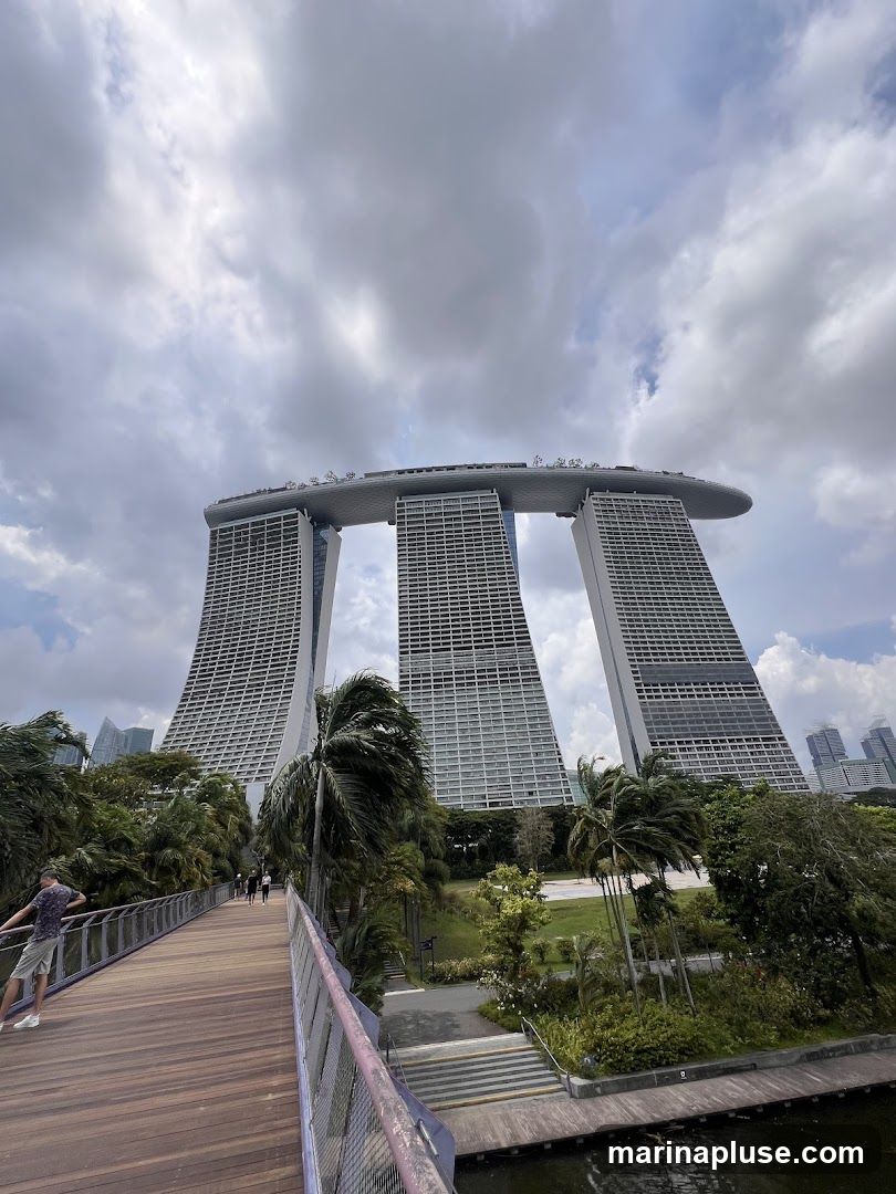 A wide-angle shot of the Marina Bay Sands hotel in Singapore on a cloudy day. The iconic three towers are connected by a large, boat-shaped structure at the top. In the foreground, a wooden walkway with a purple railing leads towards the hotel. Lush greenery and palm trees surround the walkway and the base of the hotel. A few people are visible walking on the walkway and in the park area.