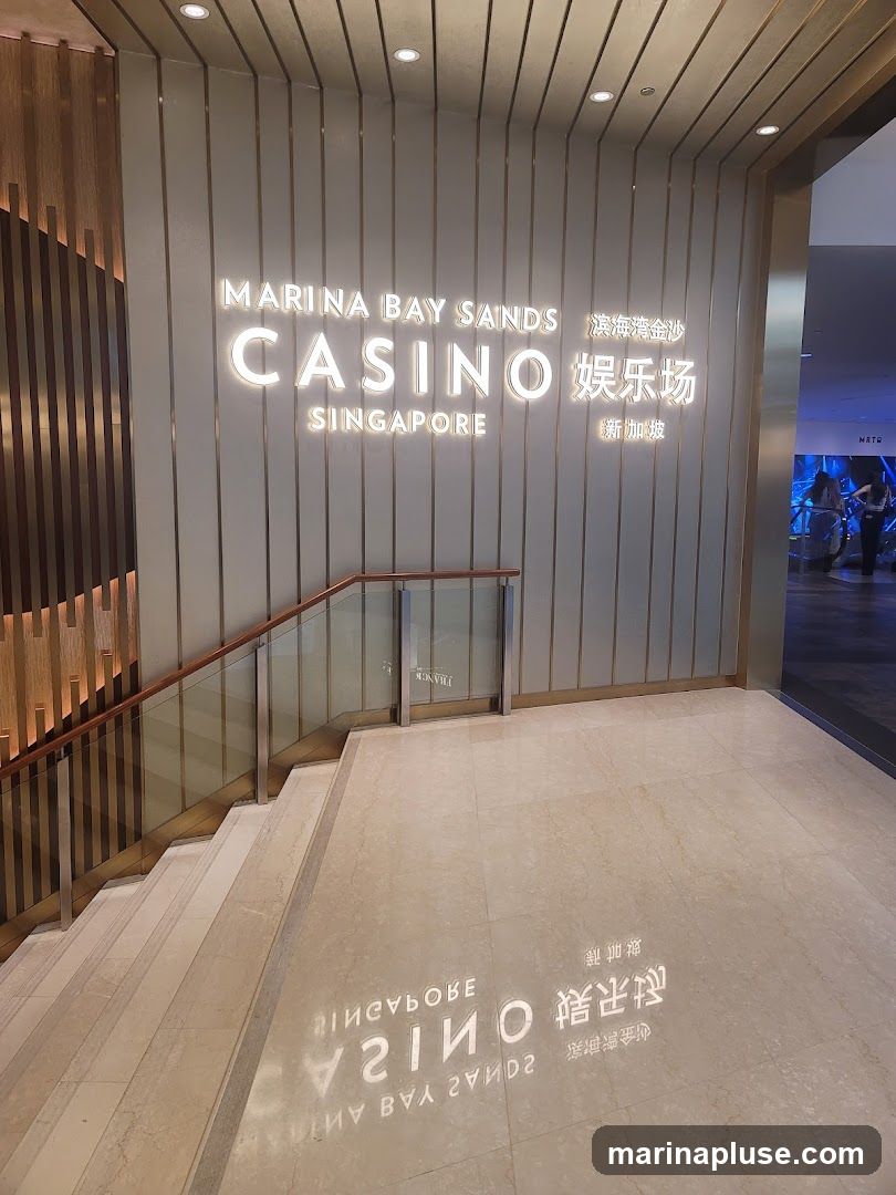 A well-lit interior hallway with a prominent sign for Marina Bay Sands Casino Singapore. The sign is illuminated with white lettering and is mounted on a wall with vertical metallic accents. To the left, a staircase with a glass railing leads upwards. The floor is made of polished marble, reflecting the signage. In the background, a glimpse of a brightly lit area with people can be seen.