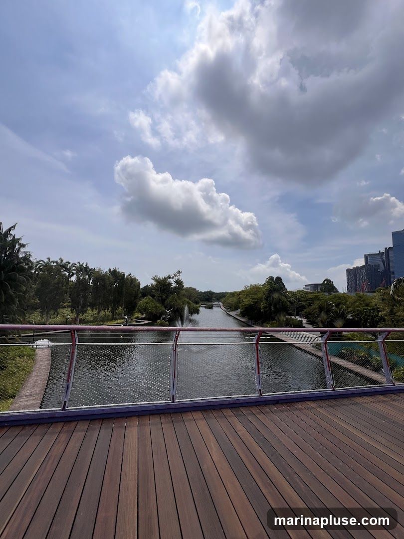 A wooden walkway with a railing overlooks a calm canal. Fountains are visible in the canal, and lush green trees and foliage line the banks. In the distance, modern buildings rise against a cloudy sky.