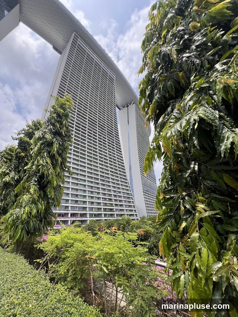 A low-angle shot of the Marina Bay Sands hotel in Singapore, with lush green trees and foliage in the foreground. The iconic three towers are visible, connected by the SkyPark at the top. The sky is partly cloudy.