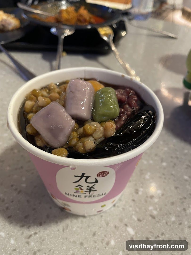 A close-up of a dessert in a pink and white branded cup. The dessert appears to be a jelly or pudding base topped with various ingredients including red beans, taro balls, green mochi, and what looks like corn or barley. The cup has a logo that reads "九 鮮 NINE FRESH" in Chinese and English. In the background, out of focus, are other dishes and utensils on a table.