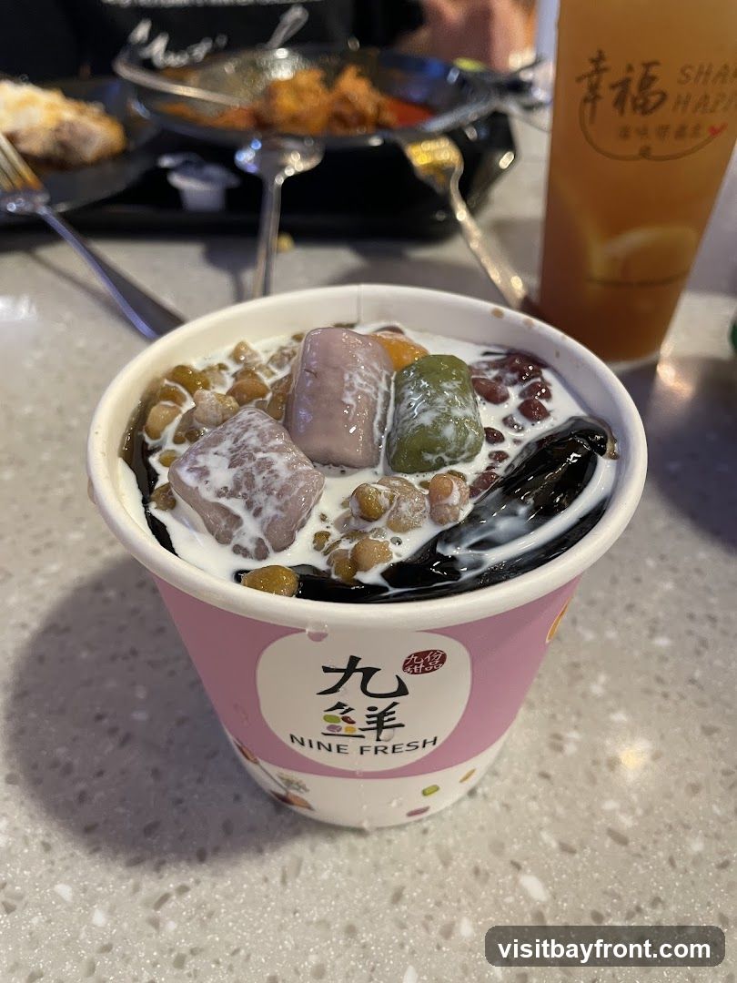 A close-up of a dessert cup filled with various toppings including taro balls, red beans, and grass jelly, all covered in a creamy liquid. The cup has a pink band with the logo "九羊 NINE FRESH" printed on it. In the background, a glass of iced tea with a logo that reads "幸福 SHAKE HAPPY" is visible, along with plates of food.