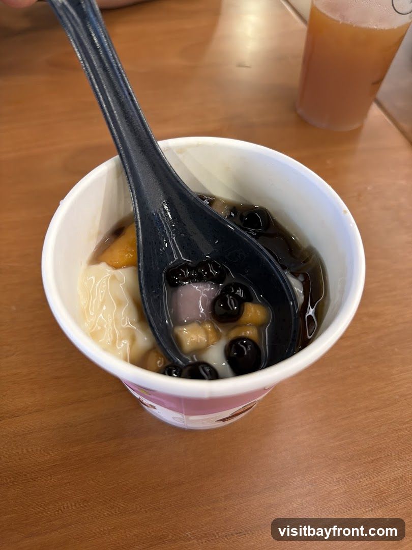 A close-up shot of a white paper cup filled with a dessert. The dessert appears to be a creamy base topped with dark syrup, boba pearls, and pieces of fruit. A black spoon is partially submerged in the dessert, scooping up some of the toppings. In the background, a blurred glass of amber-colored liquid is visible on a wooden table.