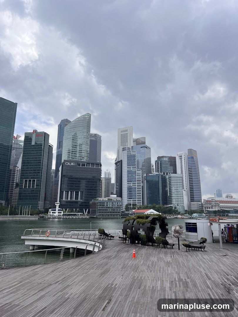 A wooden promenade with a curved walkway extends into a body of water, with a modern city skyline in the background under a cloudy sky. A unique, circular metal sculpture filled with greenery is situated on the promenade, along with several benches and an orange traffic cone. A small white kiosk or booth is also visible on the right side of the promenade.