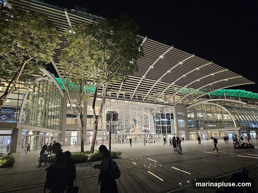 A wide shot of a large, modern building complex at night. The architecture features a distinctive curved, slatted roof illuminated with white lights, and large glass facades. Green lighting accents parts of the structure. The area in front of the building is a wide, paved plaza with people walking and gathering. Trees are planted in front of the building, and some are illuminated. The overall scene suggests a vibrant urban space, possibly a shopping mall or entertainment venue.