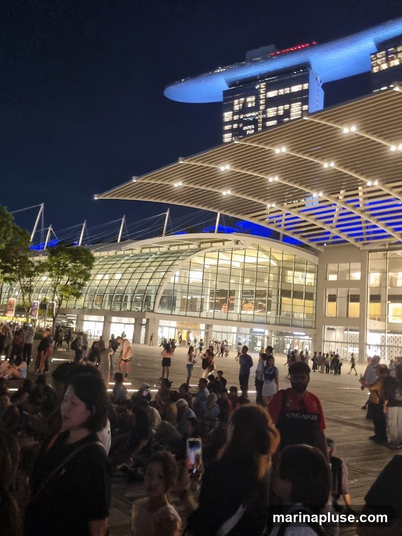 A crowd of people are gathered outdoors at night in front of a large, modern building with a glass facade. The building is illuminated, and in the background, a distinctive skyscraper with a boat-like structure on top is visible, also lit up. The atmosphere appears to be lively, with many people sitting and standing, some looking towards the building or the sky.