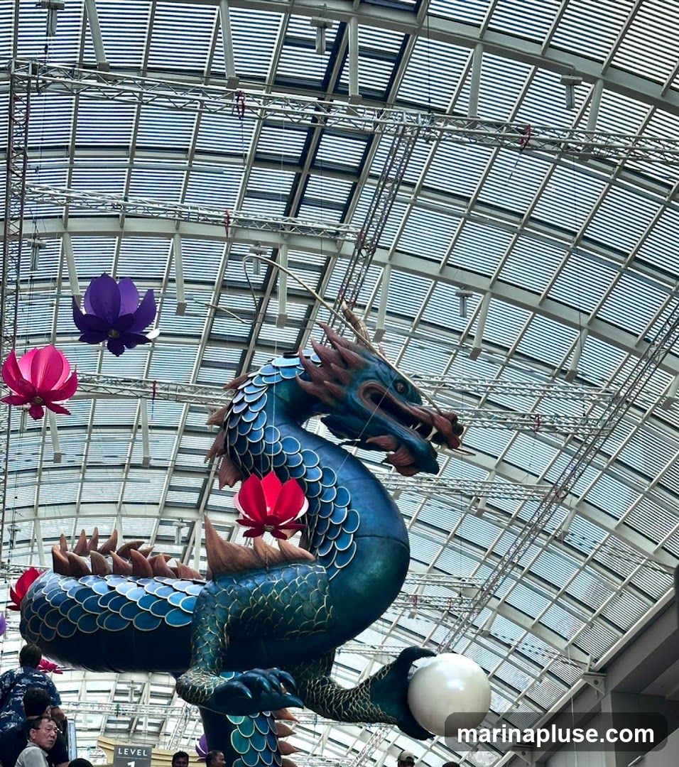 A large, blue, ornate dragon sculpture is suspended from a ceiling with a glass and metal roof. The dragon is holding a white sphere in one of its claws. Several large, colorful lotus-shaped lanterns (pink, purple, and red) are also hanging from the ceiling around the dragon. People are visible at the bottom of the image, looking up at the decoration.