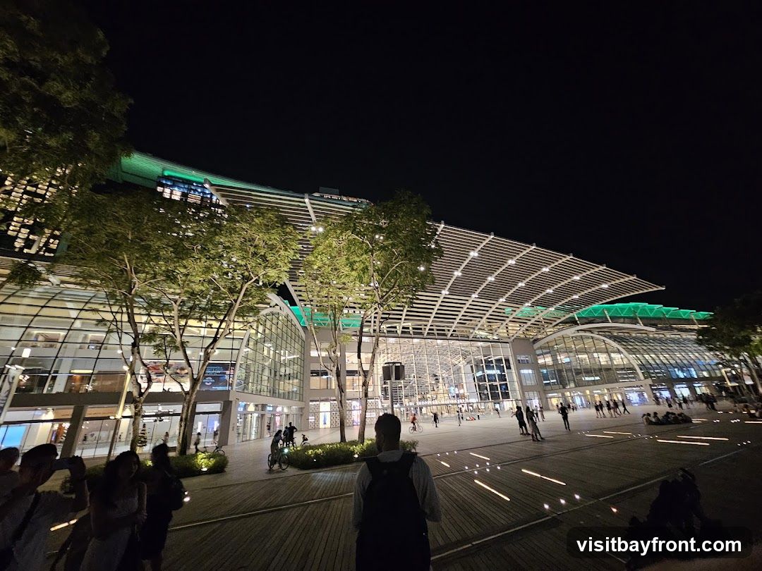A wide-angle shot of a large, modern building complex at night. The building features a distinctive curved roof structure with white latticework and green lighting accents. Large glass facades reveal multiple levels of interior spaces. Trees are strategically placed in front of the building, illuminated by soft lighting. People are visible walking around the plaza in front of the building, some with bicycles. The ground is paved with wooden planks and features embedded lights that create a pattern. The overall scene suggests a vibrant urban environment or a shopping and entertainment district.