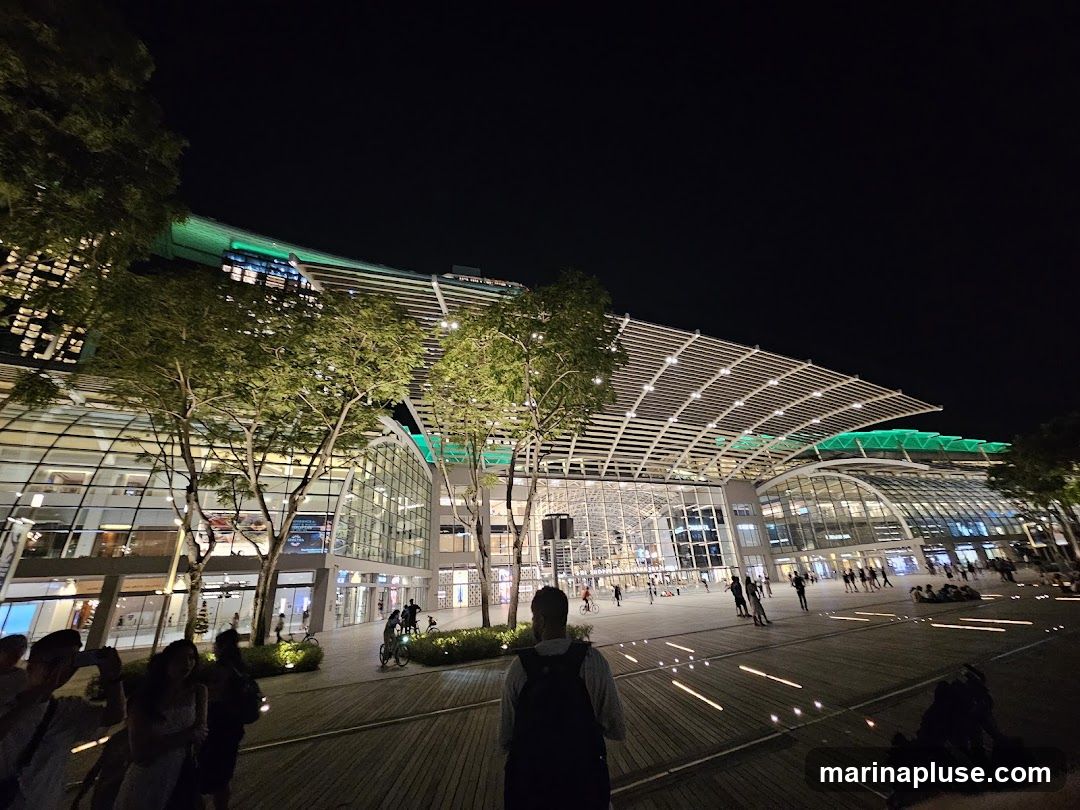 A wide-angle shot of a large, modern building complex at night. The building features a distinctive curved roof structure with white latticework and green lighting accents. Large glass facades reveal multiple levels of interior spaces. Trees are strategically placed in front of the building, illuminated by soft lighting. People are visible walking around the plaza in front of the building, some with bicycles. The ground is paved with wooden planks and features embedded lights that create a pattern. The overall scene suggests a vibrant urban environment or a shopping and entertainment district.