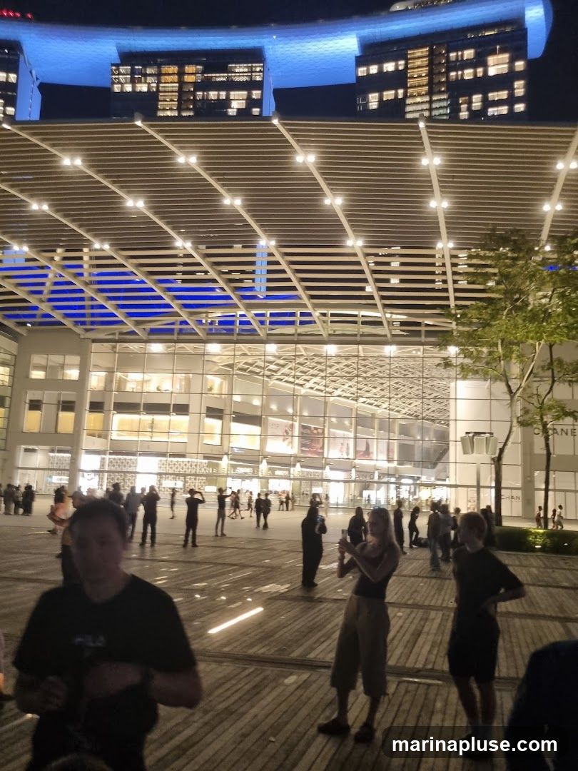 A wide shot of a modern building facade at night, illuminated by blue lights from above and white lights from within. People are visible in the foreground and on the plaza in front of the building. The architecture features a large glass-fronted section with multiple levels and a distinctive canopy structure overhead. A tree is visible to the right.