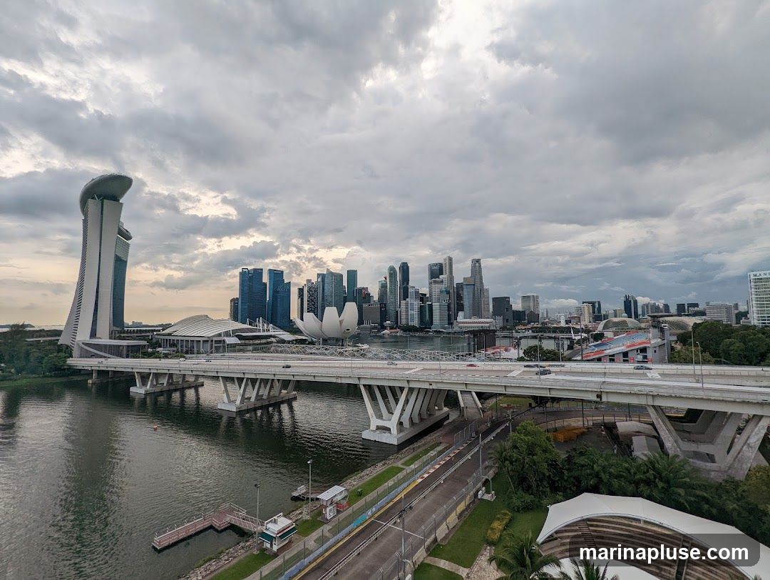 A panoramic view of the Singapore skyline under a cloudy sky. The iconic Marina Bay Sands hotel is visible on the left, with its distinctive boat-shaped top. A multi-lane bridge spans across a body of water in the foreground, with cars driving on it. The central business district's skyscrapers form the backdrop, and various other modern buildings and structures are scattered throughout the cityscape. The overall scene captures a bustling urban environment with a dramatic, overcast sky.