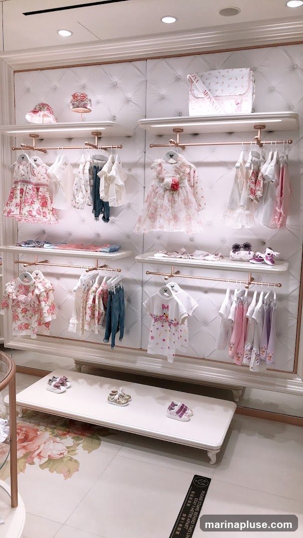 A well-lit display of children's clothing and accessories in a store. The items are neatly arranged on shelves and hanging racks against a tufted white wall. Featured are dresses, tops, jeans, hats, and shoes, many with floral patterns or cartoon characters. A diaper bag with the brand name 'MONNALISA' is visible on an upper shelf. The floor has a large floral print. A social distancing sign is visible on the floor.