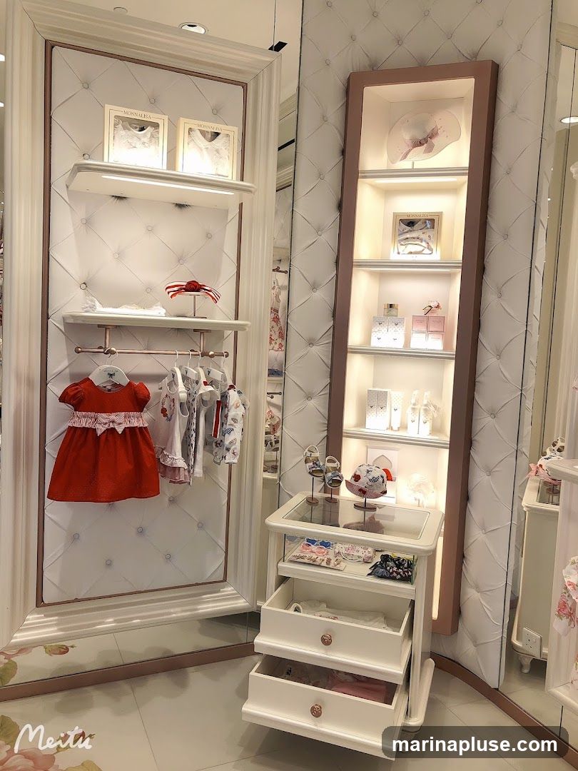 A well-lit display area in a children's clothing store. On the left, a red dress and several other garments are hung on a rack. Above them, shelves display framed items and a decorative bow. To the right, a tall display case showcases hats, perfumes, and other accessories on illuminated shelves. A rolling cart with drawers is positioned in front of the display case, holding more merchandise. The walls are upholstered with a diamond-stitched pattern.