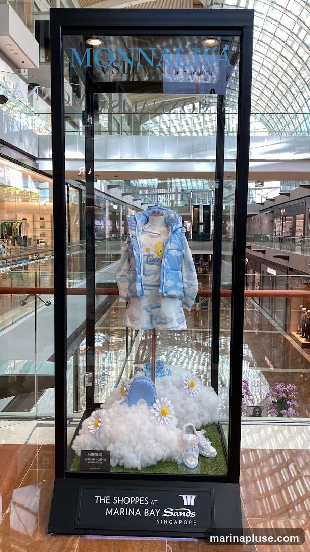 A glass display case showcases a children's outfit with a cloud and Tweety Bird theme. The outfit includes a puffer vest, long-sleeved shirt, shorts, and sneakers, all in light blue and white with cloud patterns. A small blue backpack and daisy decorations are also present. The display is set against a backdrop of a shopping mall interior. A sign at the bottom reads "THE SHOPPES AT MARINA BAY SANDS SINGAPORE".