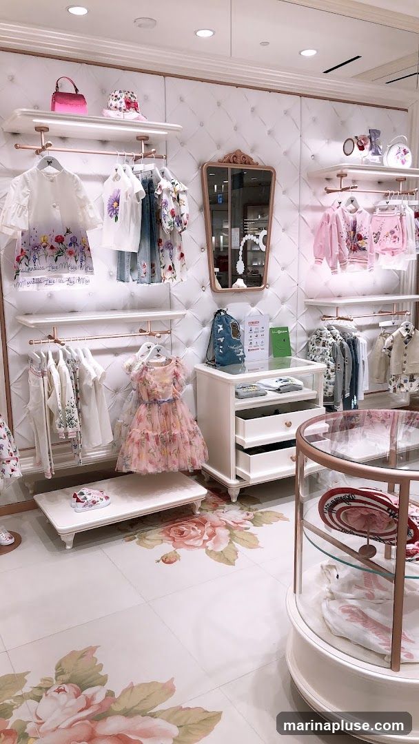 A brightly lit interior of a children's clothing store. Racks of girls' clothing, including dresses, tops, and jackets, are displayed against a white tufted wall. Accessories like hats and handbags are on shelves. A display case shows more clothing items. The floor is decorated with large floral prints.