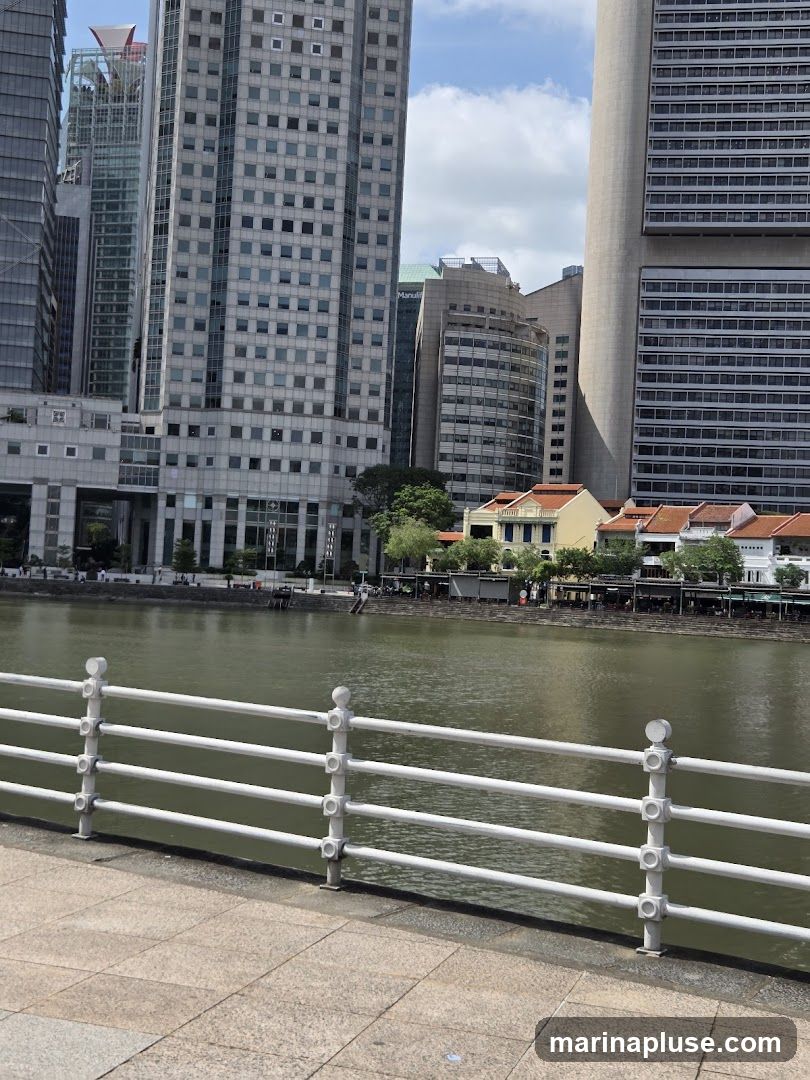 A view across a body of water towards a cityscape featuring a mix of modern skyscrapers and older, low-rise buildings with red-tiled roofs. A white railing runs along the foreground, separating a paved walkway from the water. The sky is partly cloudy.