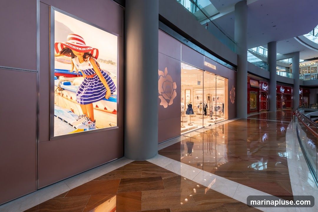 A brightly lit storefront of a children's clothing store is visible in a modern shopping mall. The store features a large glass facade displaying mannequins dressed in children's apparel. To the left, a large illuminated advertisement showcases a young girl in a striped hat and dress. The mall's polished wooden floor reflects the store lights and the surrounding architecture. Other store fronts are visible in the background.