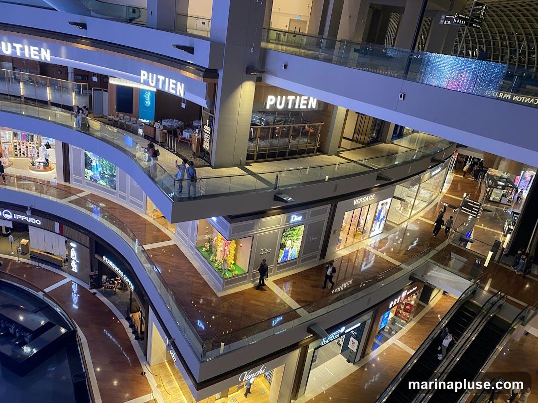 An interior view of a modern shopping mall with multiple levels, escalators, and various shops. Visible shop names include Putien, Dior, Versace, Starbucks Coffee, and Ippudo. The mall features polished wooden floors and glass railings. People are seen walking on the different levels and using the escalators.