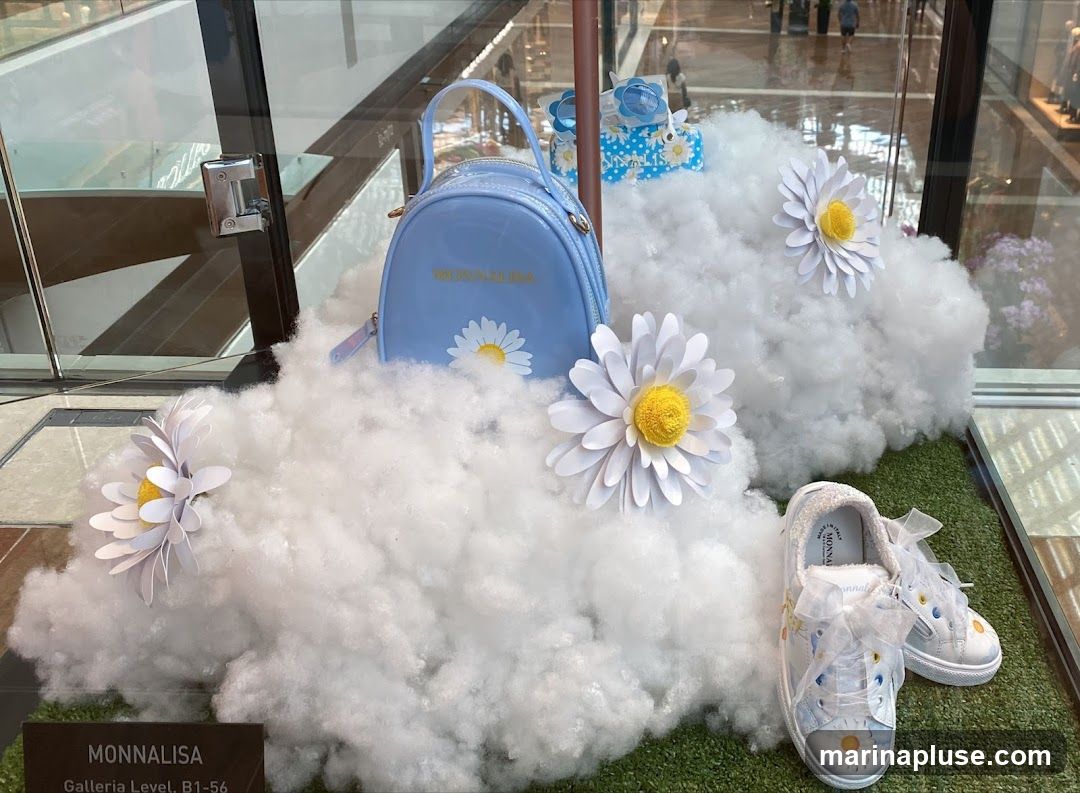 A store window display featuring a light blue patent leather backpack with a daisy design and the brand name "MONNALISA" in gold lettering. Beside it are a pair of white sneakers adorned with daisy prints and white ribbon laces. The items are placed on artificial clouds and grass, with large paper daisies as decoration. A small blue gift box with a daisy pattern is also visible. A sign at the bottom reads "MONNALISA Galleria Level, B1-56".