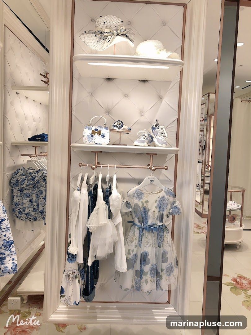 A display of children's clothing and accessories in a boutique. The display features hats, handbags, shoes, dresses, and shirts with floral patterns in blue and white. The clothing is neatly arranged on shelves and hangers against a tufted white wall.