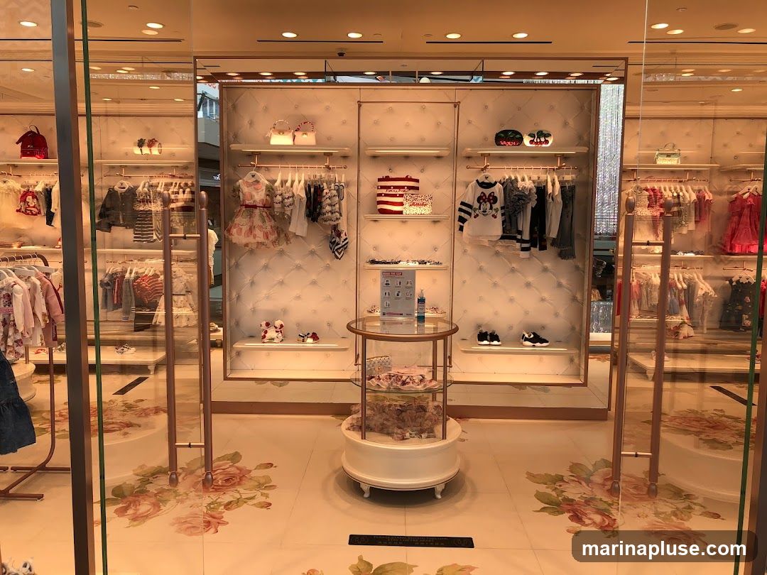 The interior of a brightly lit children's clothing store. Racks of clothing, including dresses, shirts, and pants, are displayed on shelves and hanging racks. Decorative elements like quilted panels and floral floor decals add to the aesthetic. A circular display stand in the center showcases more merchandise.