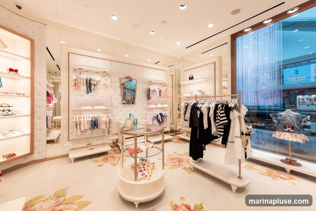 The interior of a brightly lit, elegant children's clothing boutique. Racks of dresses and outfits are displayed on white, tufted walls. A glass display case in the center showcases more children's clothing. The floor is decorated with a floral pattern. A large window on the right offers a view of another store with a mannequin dressed in a denim jacket and a tutu.