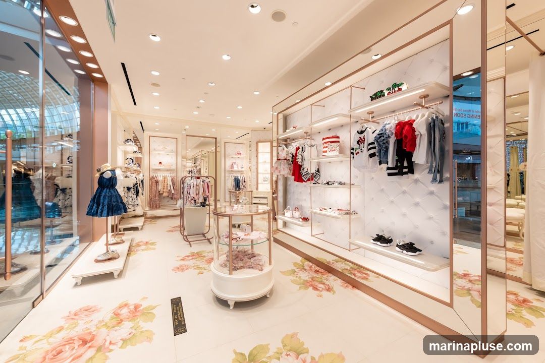 The interior of a brightly lit children's clothing store. Racks and shelves display a variety of clothing items, including dresses, shirts, and pants, in pastel colors and floral patterns. A mannequin in the foreground wears a dark blue dress. The floor is white with a large floral print. Mirrored panels are visible on the right side of the store.