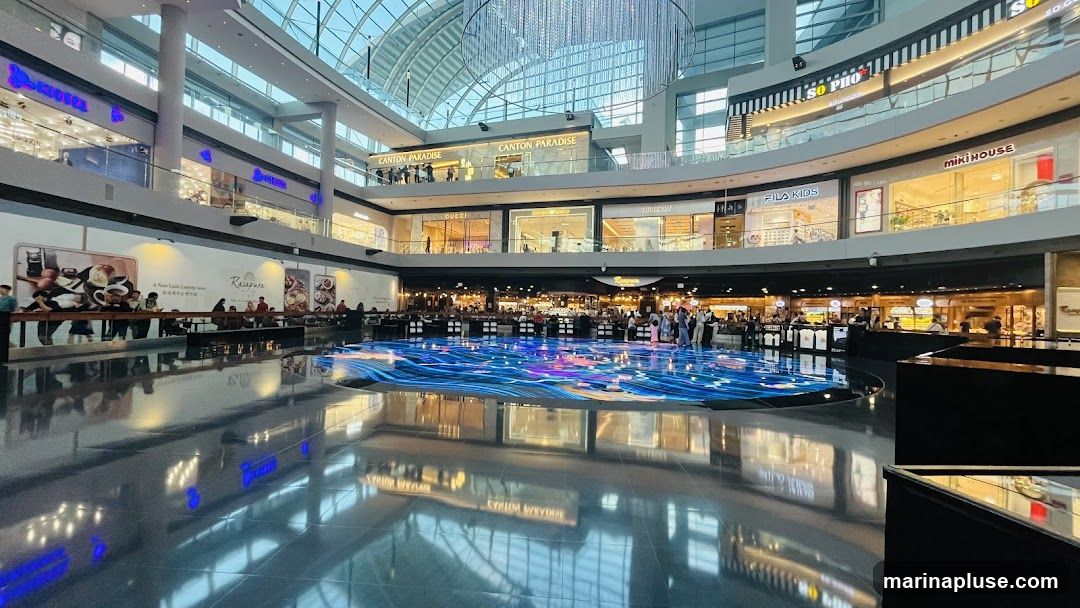 A wide shot of the interior of a modern, multi-level shopping mall. A large, circular digital display on the floor shows a dynamic, wave-like pattern of blue and purple lights. Several shops and restaurants are visible on different levels, with their signage illuminated. People are seen walking and dining in various areas. The ceiling is a large glass dome, allowing natural light to filter in, and a large, ornate chandelier hangs from the center. The polished floor reflects the lights and architecture of the mall.