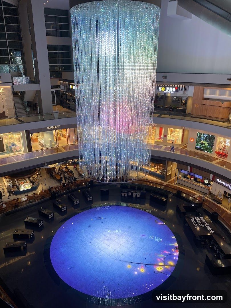 An interior view of a multi-level shopping mall atrium. A large, cylindrical chandelier made of numerous sparkling lights hangs from the ceiling, casting colorful reflections. Below, a circular digital display on the floor shows a blue, grid-like pattern with animated elements. Surrounding the atrium are various shops and dining establishments, including visible signage for Burberry, So Pho, and Ramen. People are seen walking and dining on different levels.