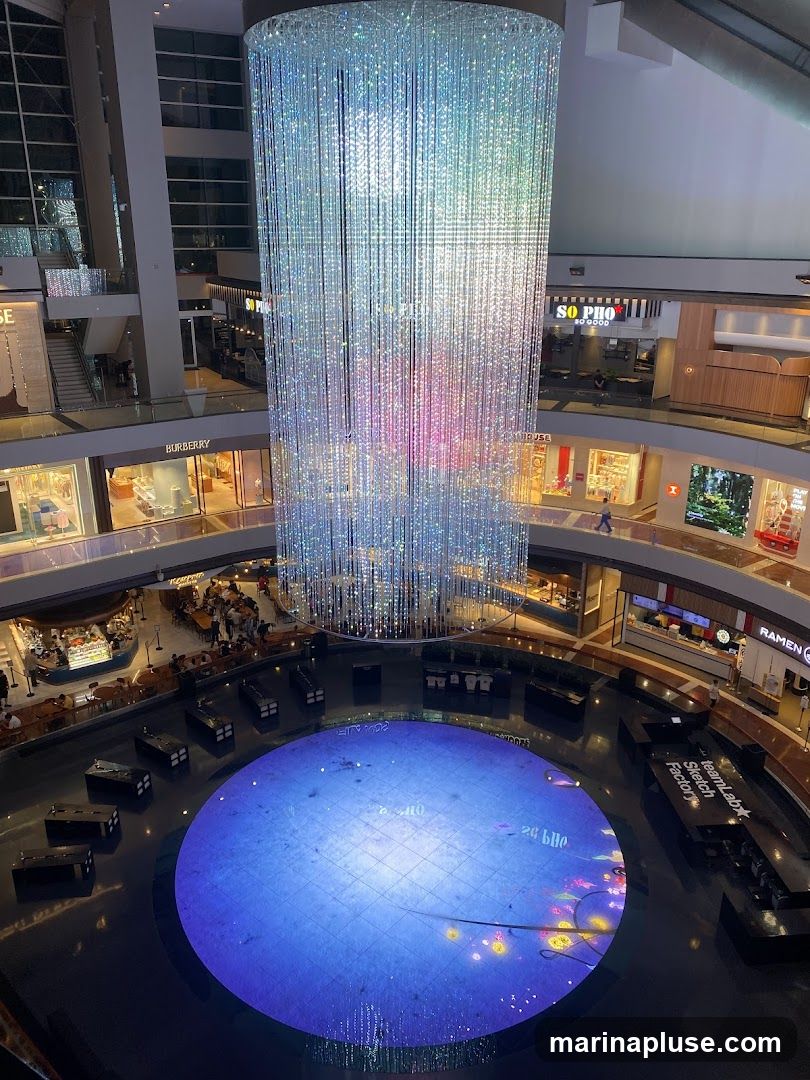 An interior view of a multi-level shopping mall atrium. A large, cylindrical chandelier made of numerous sparkling lights hangs from the ceiling, casting colorful reflections. Below, a circular digital display on the floor shows a blue, grid-like pattern with animated elements. Surrounding the atrium are various shops and dining establishments, including visible signage for Burberry, So Pho, and Ramen. People are seen walking and dining on different levels.