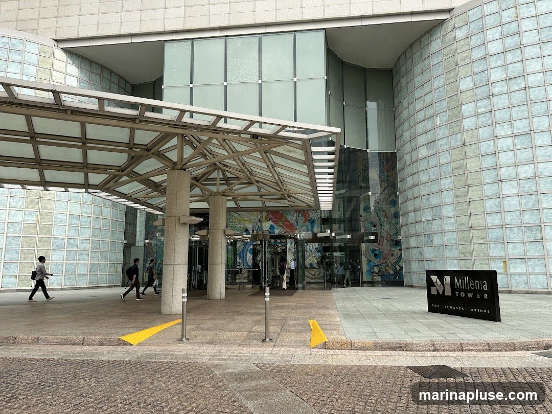 The exterior of Millenia Tower, a modern building with a glass facade and a covered entrance. A sign for "Millenia Tower" is visible on the right side of the entrance. People are walking into the building.