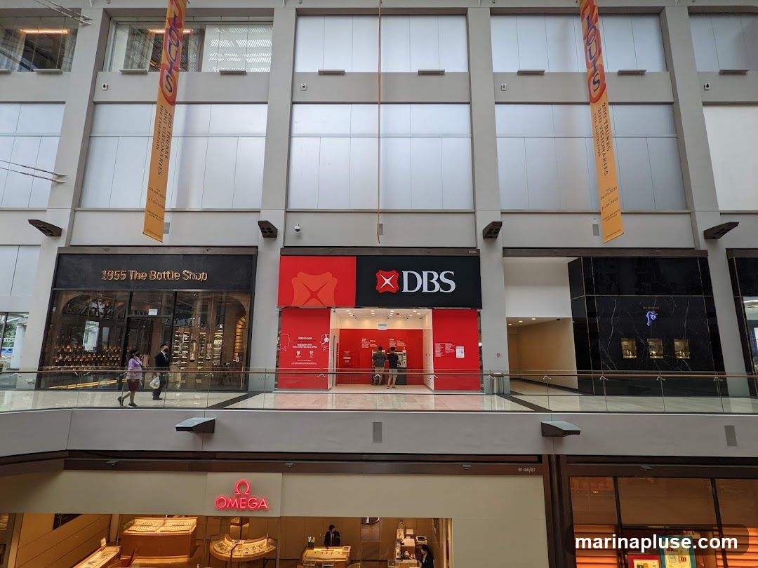 A view of a shopping mall interior showing the storefronts of DBS Bank and 1855 The Bottle Shop. The DBS bank has a red facade with the DBS logo. 1855 The Bottle Shop has a dark facade with a large display window showcasing bottles. An Omega watch store is visible on the lower level.