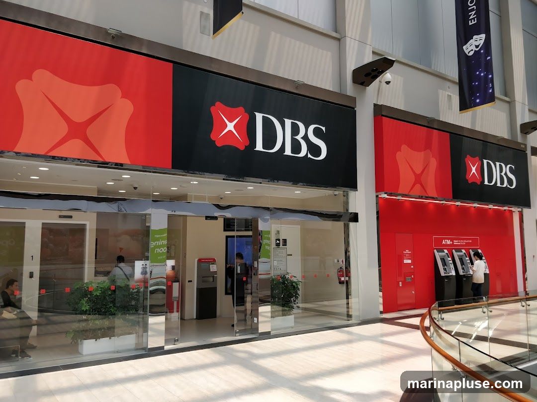 The exterior of a DBS bank branch with large red and black signage featuring the DBS logo. The entrance has glass doors and windows, with ATMs visible inside and to the right.