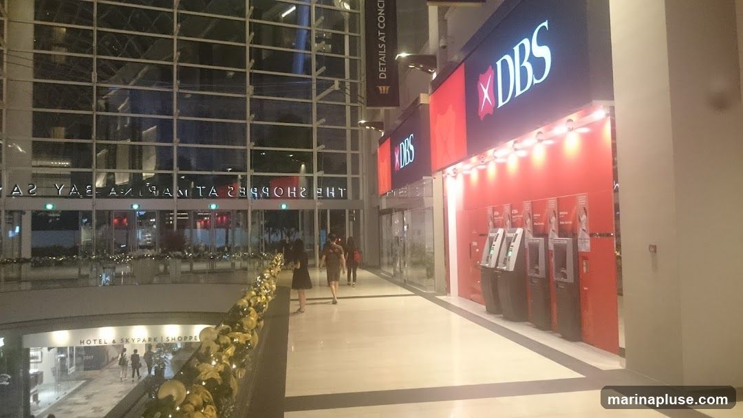 A view of a DBS bank branch entrance with ATMs visible. The bank has a prominent red facade with the DBS logo displayed. The entrance is located within a modern shopping mall, with reflections of the mall's interior visible on the glass facade.