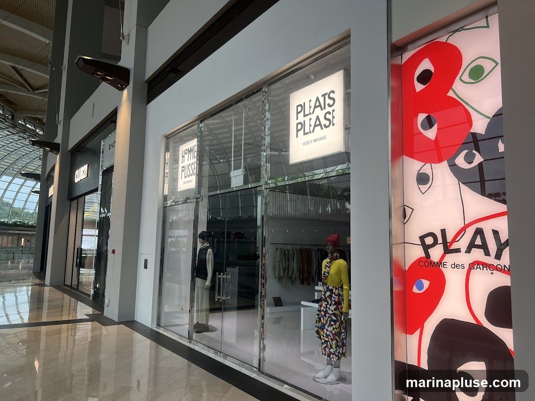 The exterior of a retail store with large glass windows and doors. A sign above the entrance reads "PLEATS PLEASE ISSEY MIYAKE". Inside, mannequins are displayed wearing clothing. To the right, a large graphic display features the text "PLAY COMME des GARÇONS" with abstract eye-like shapes.