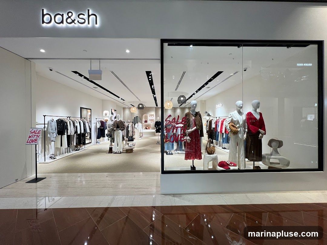 The exterior of a Ba&sh clothing store. The storefront features a large glass window displaying mannequins dressed in fashionable outfits. Inside, racks of clothing are visible, along with more mannequins. A sign that reads "SALE WITH LOVE" is placed near the entrance. The store's name, "ba&sh", is prominently displayed above the entrance.