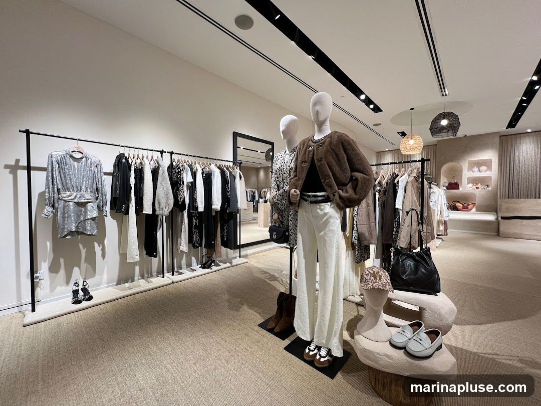 A well-lit interior of a fashion boutique, showcasing clothing on racks and mannequins. A silver sequined dress hangs on a rack, alongside various tops, jackets, and pants in neutral tones. Two mannequins are displayed, one wearing a patterned dress and the other a brown furry jacket with white pants. Accessories like handbags, hats, and shoes are also visible. The store features modern lighting and decor, with shelves displaying handbags in the background.