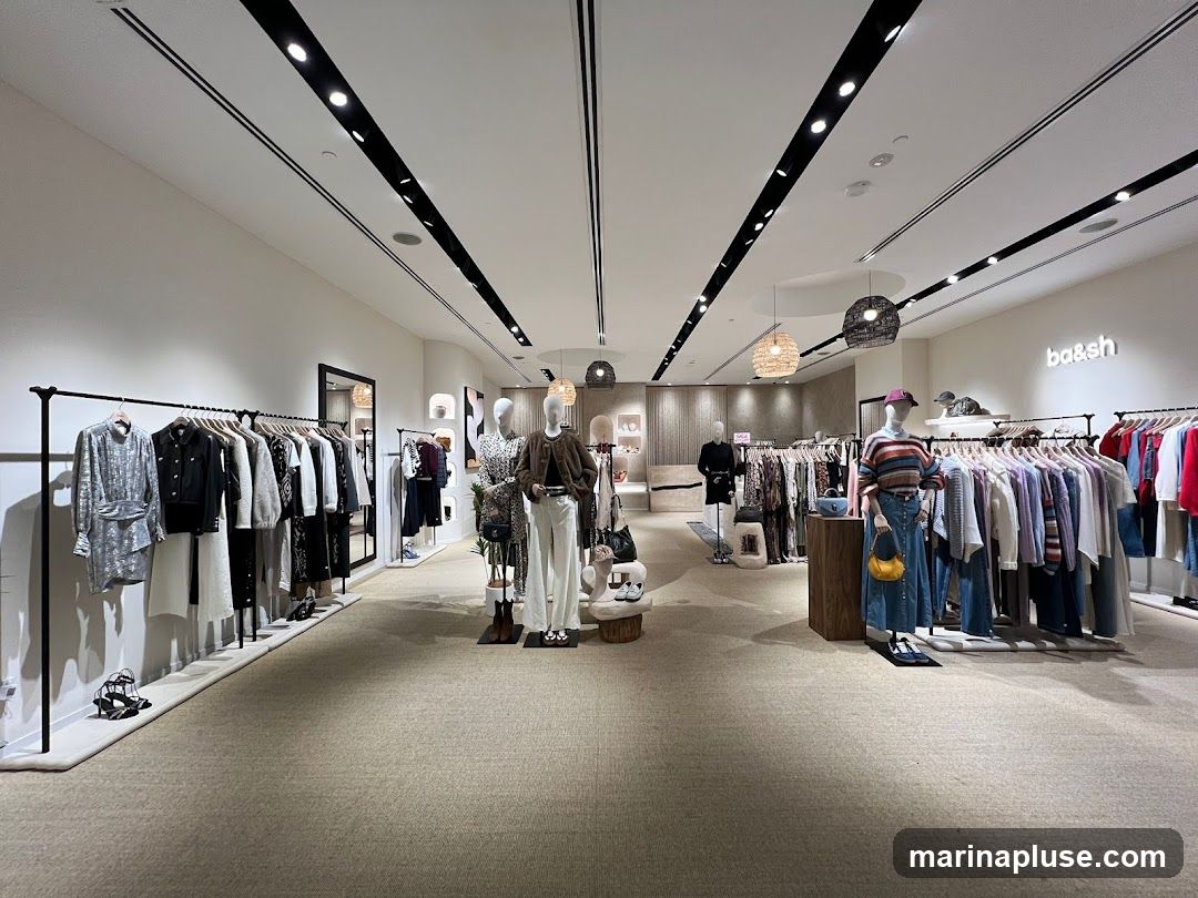 The image shows the interior of a clothing store. Racks of clothing are displayed on the left and right sides of the store. Mannequins are placed throughout the store, showcasing outfits. The store has a modern and clean aesthetic with white walls and a neutral-colored carpet. The brand name 'ba&sh' is visible on the back wall.