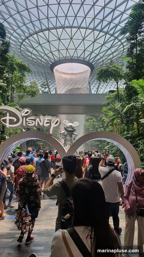 A crowd of people are gathered in front of a large archway with the Disney logo and a statue of Mickey Mouse. Behind the archway, a large indoor waterfall cascades down from a geodesic dome ceiling. Lush greenery surrounds the area, creating a tropical atmosphere.