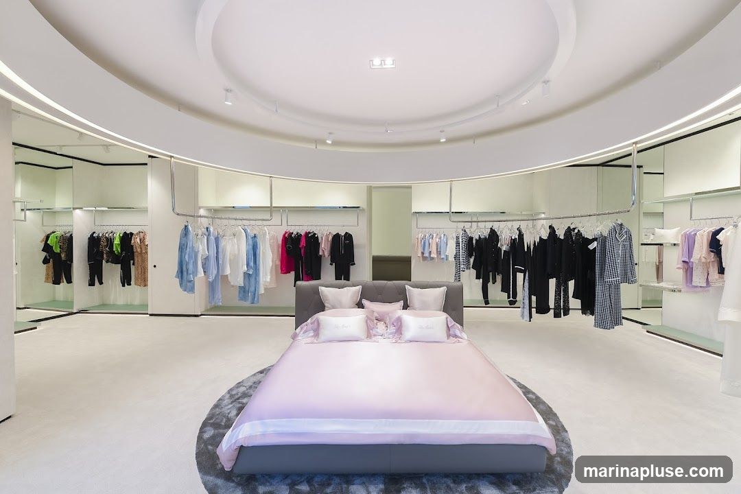 A modern retail space designed to resemble a bedroom, featuring a bed with pink bedding as a central display. Racks of clothing are arranged around the perimeter of the circular room, with various styles and colors of apparel visible. The lighting is soft and ambient, creating a luxurious atmosphere.